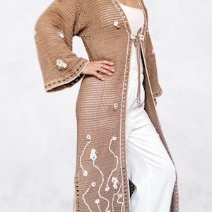 May include: A long, brown crocheted cardigan with white crocheted flowers and a tie at the waist.