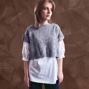 May include: A woman wearing a gray crocheted top over a white shirt and black pants. The top has a square pattern and is cropped.
