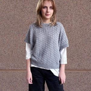 May include: A gray knitted sweater with a delicate openwork pattern. The sweater has a round neckline and short sleeves. The model is wearing the sweater over a white shirt.