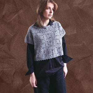 Könnte beinhalten: Eine Frau trägt ein graues Häkeltop über einem schwarzen Langarmshirt und einer schwarzen Hose. Das Top hat einen gewellten Saum und eine lockere, fließende Passform.