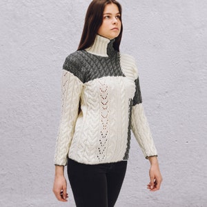 Könnte beinhalten: Eine Frau trägt einen weißen und grauen Strickpullover mit Rollkragen. Der Pullover hat ein Zopfmuster und ein kontrastierendes graues Panel auf der Schulter.