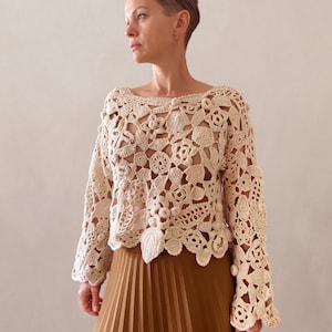 May include: A cream-colored crocheted top with a floral design and bell sleeves. The top is loose-fitting and has a scalloped hem.