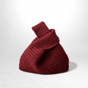 May include: A burgundy-colored, knitted bag with a unique, folded design. The bag is made of a textured material and has a structured shape. The top portion is folded over, creating a distinctive look. The bag is set against a white background.