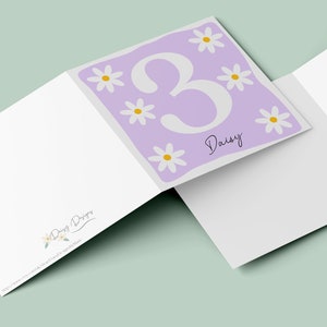 Pode incluir: Um cart&atilde;o roxo com um n&uacute;mero 3 branco e o nome Daisy escrito em it&aacute;lico. O cart&atilde;o &eacute; decorado com margaridas brancas com centros amarelos.