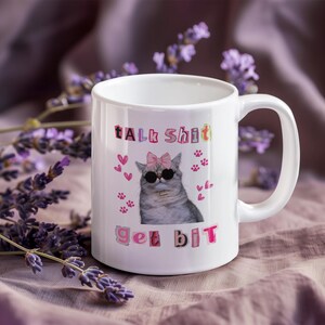 May include: White ceramic mug featuring a cat wearing sunglasses and a pink bow. The mug has the text "Talk Shit Get Bit" in colorful block letters, along with pink hearts and paw prints. The mug has a curved handle.