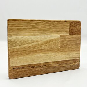 Puede incluir: Una tabla de cortar rectangular de madera de roble marrón claro. La tabla tiene un patrón de veta de madera natural y está hecha de varias piezas de madera unidas.