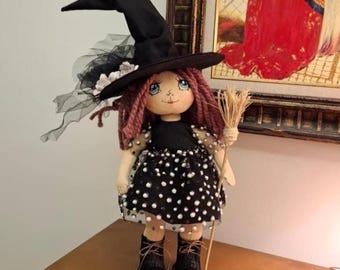 May include: A handmade doll dressed as a witch, featuring a black pointed hat, a black dress with white polka dots, and black boots. The doll has red yarn hair and holds a small broom. The doll is standing on a wooden surface.