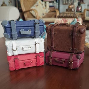 Peut inclure: Quatre petites valises empilées sur une surface en bois. Les valises sont en cuir et ont un aspect vintage. La valise du haut est marron, la deuxième est blanche, la troisième est rouge et la valise du bas est bleue.