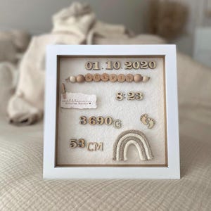 Op de afbeelding: Een witte schaduwbox met houten versieringen. De doos bevat de datum 01.10.2020, de naam "KELVIN", tijd 8:23, gewicht 3690g en lengte 53cm. Een kleine gebreide regenboog en babyvoetjes zijn ook inbegrepen.