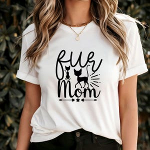 Puede incluir: Camiseta blanca con el texto "Fur Mom" en fuente negra, con dos siluetas de gatos. El diseño incluye flechas y estrellas. La camiseta tiene mangas cortas y cuello redondo.