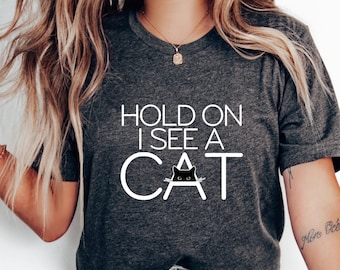 May include: A gray t-shirt with the text "Hold on I see a cat" in white letters. A black cat silhouette is inside the word "cat".