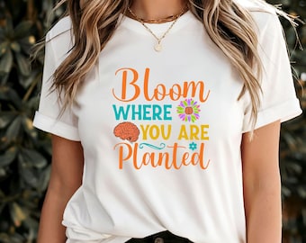 Puede incluir: Camiseta blanca con el texto "Bloom where you are planted" en naranja, amarillo y azul. Se incluye un gráfico de cerebro y flor.
