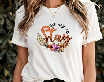 Puede incluir: Camiseta blanca con un diseño gráfico que dice "One More Day Stay" en estampado de leopardo naranja y negro con un diseño floral alrededor del texto.