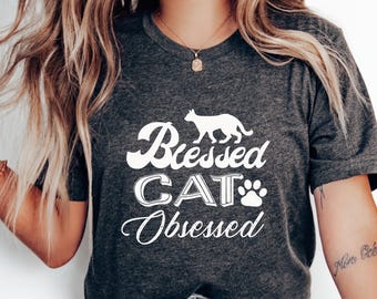 Puede incluir: Camiseta gris oscuro con las palabras blancas "Blessed CAT Obsessed" y una silueta de gato. La camiseta también presenta una huella de pata de gato. Una camiseta informal para los amantes de los gatos.