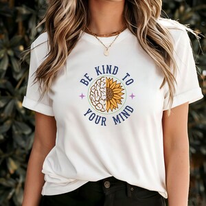 May include: White t-shirt with a blue and green circle graphic that says "Be Kind To Your Mind". The graphic features a brain and a sunflower.