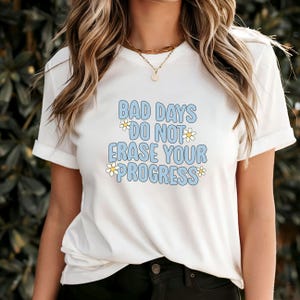 May include: White t-shirt featuring the text "BAD DAYS DO NOT ERASE YOUR PROGRESS" in light blue, with a darker blue outline and daisy accents. The shirt has short sleeves and a round neck.