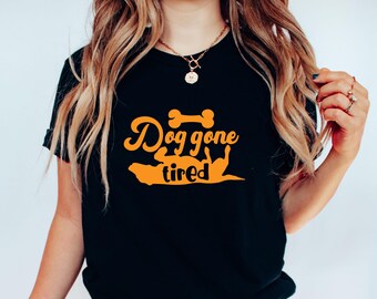 Op de afbeelding: Zwart T-shirt met een oranje afbeelding met de tekst "Dog gone tired" met een silhouet van een hond en een bot.