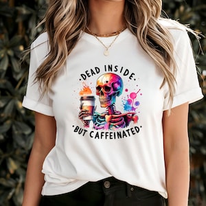 Pode incluir: Camiseta branca com um design colorido de esqueleto segurando uma xícara de café. O design inclui o texto "DEAD INSIDE. BUT CAFFEINATED." O esqueleto é retratado com tons vibrantes de arco-íris e respingos em aquarela.