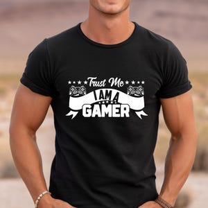 May include: Black t-shirt with white text that reads "Trust Me I Am A Gamer" with a graphic of two video game controllers.