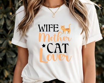 Puede incluir: Camiseta blanca con las palabras "WIFE Mother *CAT Lover" en letras negras y melocotón. Un gato de dibujos animados está encima de la palabra "Mother". La camiseta tiene las mangas enrolladas.