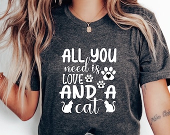 Peut inclure: Un t-shirt gris avec le texte "All you need is love and a cat" en blanc. Le texte est entouré d'empreintes de pattes et de silhouettes de chats.