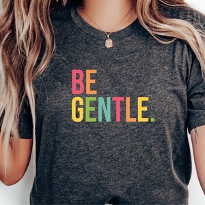 Könnte beinhalten: Ein dunkelgraues T-Shirt mit dem Aufdruck "BE GENTLE." in Regenbogenfarben.
