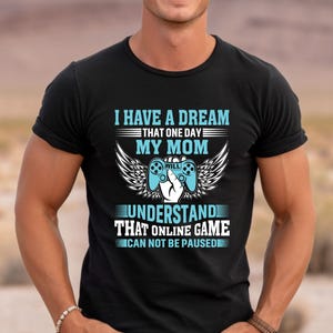 May include: Black t-shirt with a gaming-themed design. The text reads "I HAVE A DREAM THAT ONE DAY MY MOM WILL UNDERSTAND THAT ONLINE GAME CAN NOT BE PAUSED" in white and blue, with a controller and wings graphic.