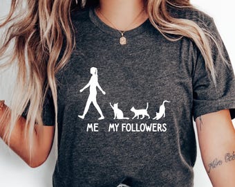 Może przedstawiać: Szary t-shirt z białym nadrukiem przedstawiającym osobę idącą z czterema kotami za sobą. Pod nadrukiem znajduje się napis "ME MY FOLLOWERS".