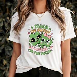 May include: White t-shirt featuring a colorful graphic with the text "I TEACH THE CUTEST CLOVERS IN THE PATCH." The design includes two cartoon clovers with sunglasses and a pink bow, surrounded by hearts and shamrocks.