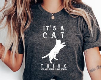 May include: A dark gray t-shirt with a white graphic that says "It's a cat thing you wouldn't understand". The graphic includes a white silhouette of a cat jumping.