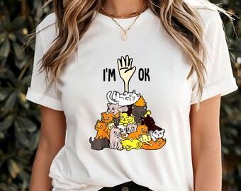 May include: White t-shirt with a graphic of a hand with a pile of cats on top of it. The text "I'm OK" is printed on the shirt.
