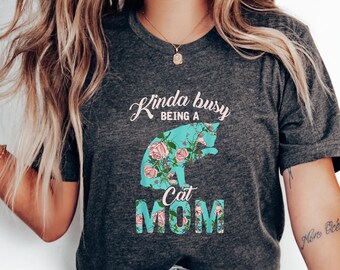 May include: A gray t-shirt with the text "Kinda busy being a Cat Mom" in white and blue lettering. The text is surrounded by a floral design with pink roses and green leaves. The shirt is being worn by a person with long blonde hair.
