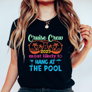 May include: Black t-shirt with a colorful graphic that reads "Cruise Crew 2025 most likely to HANG AT THE POOL." The design includes sunglasses, palm trees, and a cocktail. The shirt is a casual wear item.