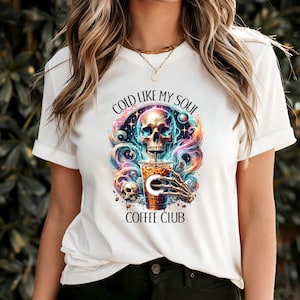 Può includere: T-shirt bianco con grafica colorata raffigurante un teschio, una tazza di caffè e una mano scheletrica. Il testo "COLD LIKE MY SOUL" è sopra il teschio e "COFFEE CLUB" sotto.