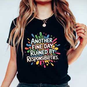 May include: Black t-shirt with the phrase "ANOTHER FINE DAY RUINED BY RESPONSIBILITIES" in a colorful splash design. The text is in a bold, playful font, with a mix of colors including blue, green, yellow, and pink.