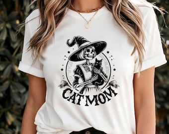 May include: White t-shirt featuring a black and white graphic of a skeleton witch holding a black cat. The design is encircled with stars and the words "CAT MOM". The shirt has short sleeves and a crew neck.