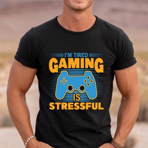 Puede incluir: Camiseta negra con un gráfico azul y naranja de un mando de videojuego. El texto "I'm Tired Gaming Is Stressful" está impreso en la camiseta.