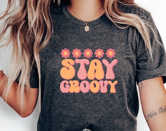 Könnte beinhalten: Ein dunkelgraues T-Shirt mit einer pinkfarbenen und orangenen Grafik, auf der "STAY GROOVY" steht, mit fünf pinkfarbenen Blumen über dem Text.