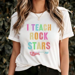 Könnte beinhalten: Weißes T-Shirt mit dem Text "I TEACH ROCK STARS" in bunten, verwaschenen Buchstaben. Eine Gitarrengrafik befindet sich unter dem Text. Das Shirt hat kurze Ärmel und einen Rundhalsausschnitt.