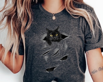 Può includere: Una maglietta grigio scuro con un gatto nero che spunta da un buco strappato nel tessuto. Il gatto ha gli occhi gialli e un'espressione seria.