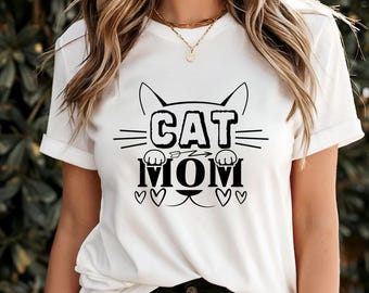 Peut inclure: T-shirt blanc avec un motif noir « CAT MOM ». Le motif comprend des oreilles de chat, des moustaches, des pattes, une flèche et des cœurs. Le t-shirt a des manches courtes et un col rond.