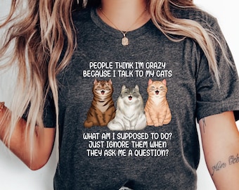 May include: A dark gray t-shirt with a humorous saying about talking to cats. The text reads "People think I'm crazy because I talk to my cats. What am I supposed to do? Just ignore them when they ask me a question?" and features three cartoon cats.