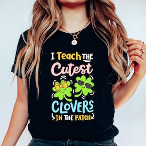 Op de afbeelding: Zwart T-shirt met de tekst "I Teach the Cutest Clovers in the Patch". Het ontwerp toont twee groene klavers, één met een gele strik en de andere met een zonnebril. De tekst is in geel, roze en blauw.