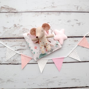 May include: A handmade beige mouse doll with a polka-dot outfit rests on a floral pillow next to a pink star. A string of pink and gray triangle flags decorates the scene, all set against a white wooden background.
