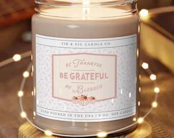 May include: A brown glass candle jar with a white label that reads "FIR & FIG CANDLE CO." and "BE THANKFUL BE GRATEFUL BE BLESSED". The candle is surrounded by white lights and is sitting on a wooden surface.