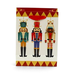 May include: A rectangular gift bag with a cream background featuring three nutcracker figures in green, red, and beige uniforms. The bag has a red ribbon handle and a decorative border of red and gold triangles at the top and bottom edges.