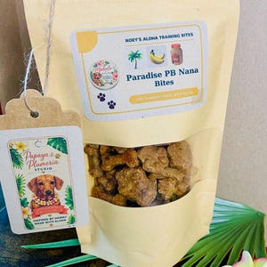 May include: A tan stand-up pouch containing brown dog treats, labeled Noey's Aloha Training Bites Paradise PB Nana Bites with icons of a palm tree, bananas, and a jar. A tag attached with twine features a dachshund illustration and the text Papaya & Plumeria Studio.