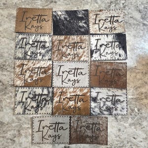 Pode incluir: Uma coleção de patches de couro com o texto "Iretta Kays" escrito à mão. Os patches apresentam vários padrões de pele de vaca em tons de castanho, preto e branco. Os patches retangulares têm bordas costuradas.