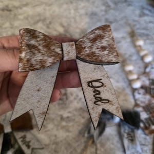 Puede incluir: Un lazo de madera con un patrón de piel de vaca en el lazo y un acabado gris en las colas. La palabra "Dad" está grabada en una cola. El lazo es un artículo decorativo.