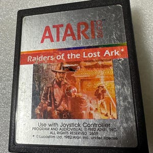 Peut inclure: Une cartouche de jeu Atari 2600 argentée avec une étiquette noire. L'étiquette présente le texte "ATARI" en blanc, suivi du texte "Raiders of the Lost Ark*" en blanc. L'étiquette comprend également un graphique d'Indiana Jones, un personnage du film Les Aventuriers de l'arche perdue, debout devant un bâtiment. L'étiquette comprend le texte "Use with Joystick Controller", "PROGRAM AND AUDIOVISUAL © 1982 ATARI, INC.", "ALL RIGHTS RESERVED 2659", et "*Lucasfilm Ltd. 1982/Atari Inc. under license".